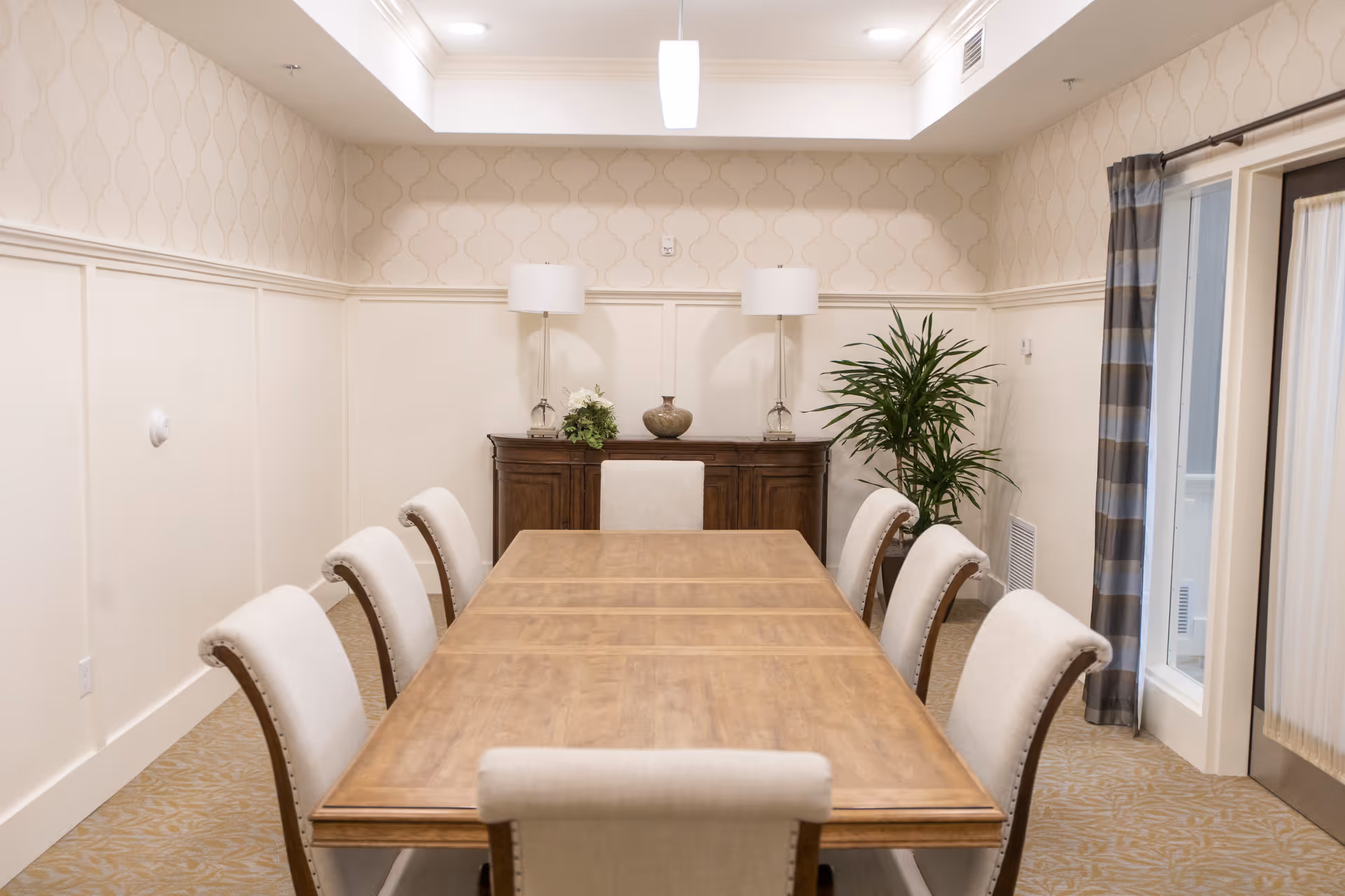 Long wooden dining table surrounded by upholstered chairs in a softly lit dining/meeting room with a sideboard, lamps, and a potted plant.