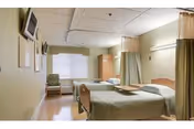 A shared skilled nursing room with two beds separated by privacy curtains, a wall-mounted TV, a window with blinds, a chair, and bedside furniture.