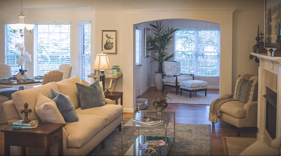 Sunlit living room with beige sofas, throw pillows, glass coffee table, fireplace and an adjacent dining area by large windows.