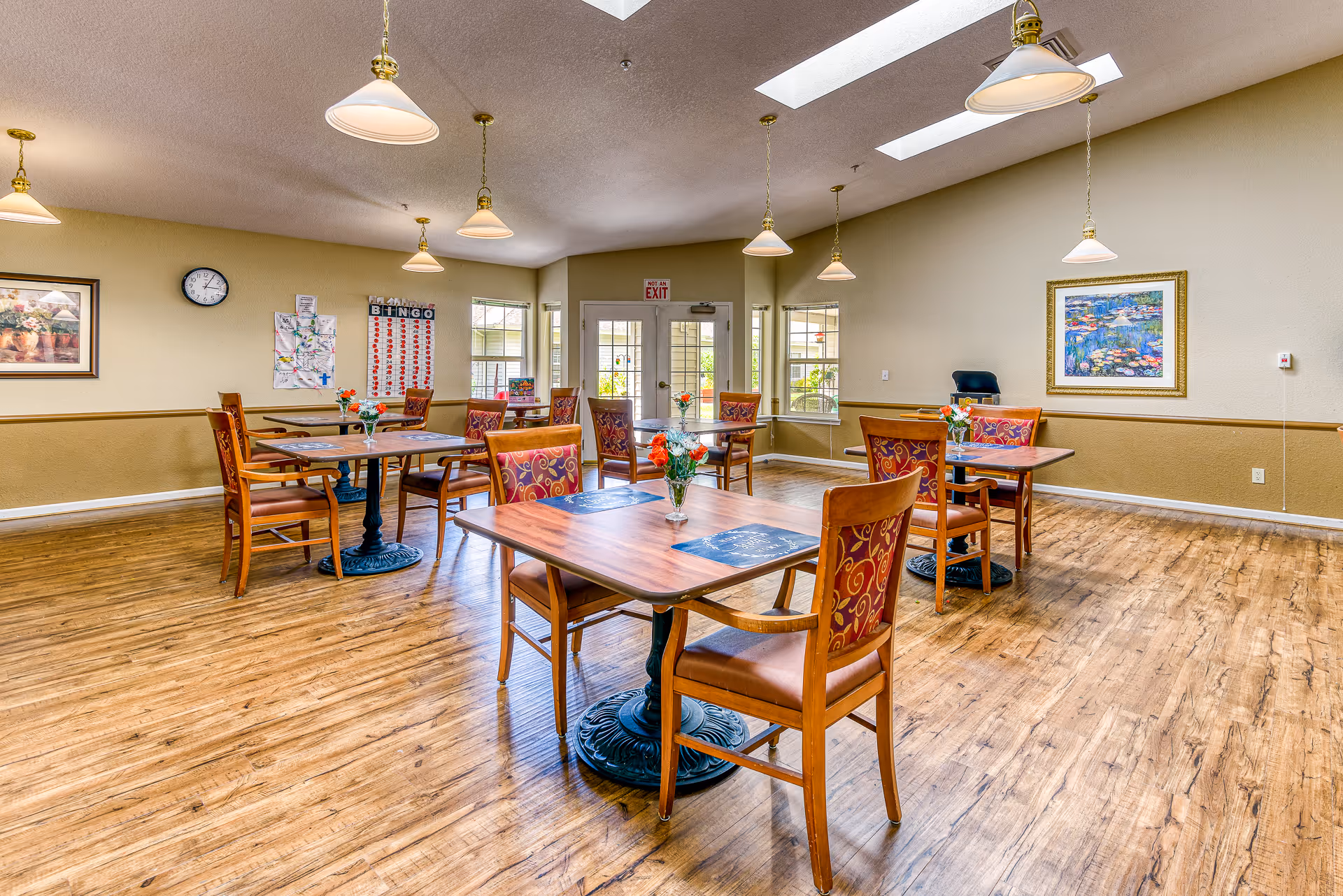 A bright and spacious dining room with wooden floors and several square wooden tables, each with four chairs featuring patterned upholstery. Each table has a small vase with flowers. The room has multiple hanging pendant lights and skylights, with windows and a door letting in natural light. Walls are decorated with framed artwork and a bingo board.