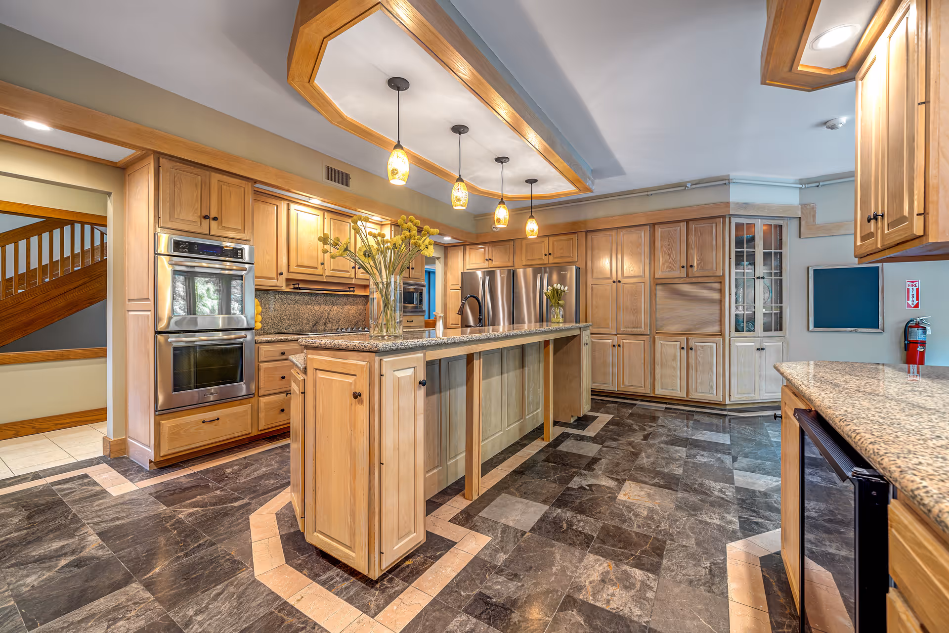 Spacious kitchen with wooden cabinets, granite countertops, and a large island in the center. Four pendant lights hang above the island, which has a vase with yellow flowers. Stainless steel double ovens and a refrigerator are visible. The floor is tiled with a dark and light pattern, and there is a fire extinguisher mounted on the wall.