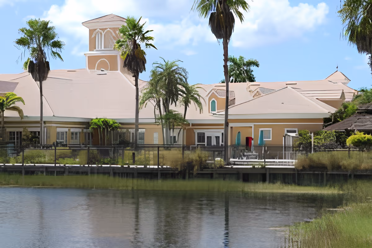 Exterior view of a senior living facility building with beige walls and a light-colored roof, surrounded by palm trees and greenery, with a body of water in the foreground reflecting the building and trees under a blue sky with some clouds.