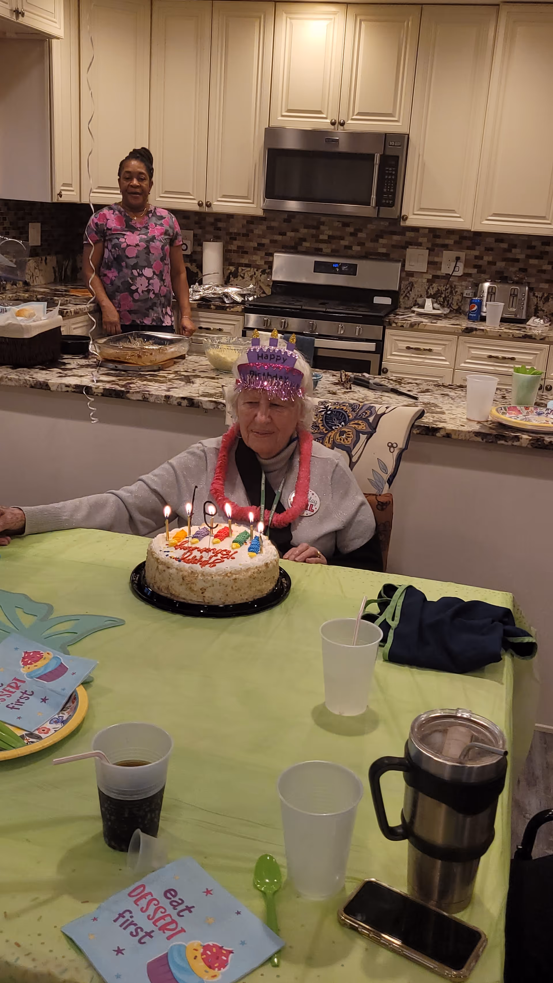 An elderly woman wearing a birthday tiara and a pink lei sits at a table with a birthday cake lit with candles in front of her. Behind her is a kitchen with a woman standing near the counter, which has various dishes and food items. The table is covered with a green tablecloth and has cups, napkins, a spoon, a travel mug, and a smartphone on it.