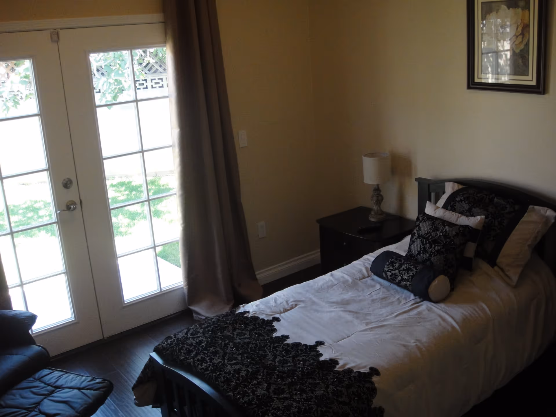 A small bedroom with a single bed dressed in patterned pillows and a nightstand with a lamp, next to French doors leading outside.
