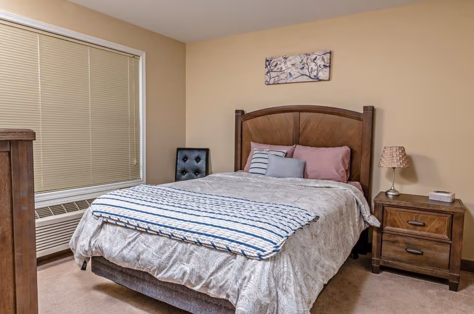 Neatly made bedroom with a wooden bed and headboard, nightstand with lamp, window with blinds, and a small chair.