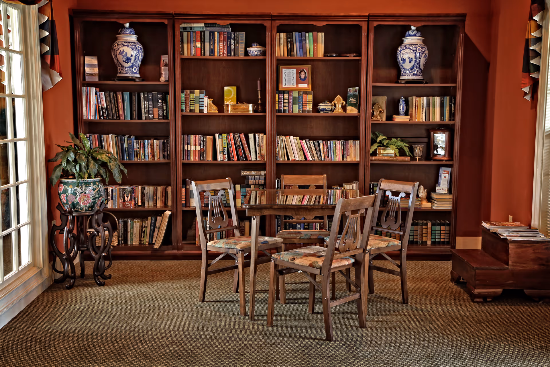 A cozy reading or sitting area with a wooden table and four chairs with patterned cushions. Behind the table is a large wooden bookshelf filled with books, decorative vases, and small plants. To the left, there is a potted plant on a decorative stand near a window with curtains. The room has warm, earthy tones with carpeted flooring.