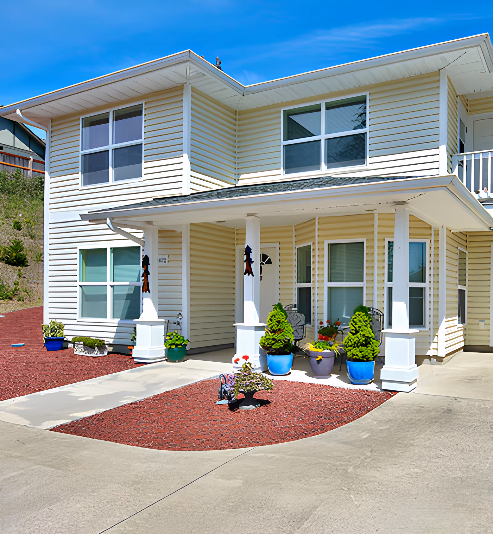 Front entrance of a two-story light-yellow siding building with a covered porch, potted plants, and a concrete driveway.