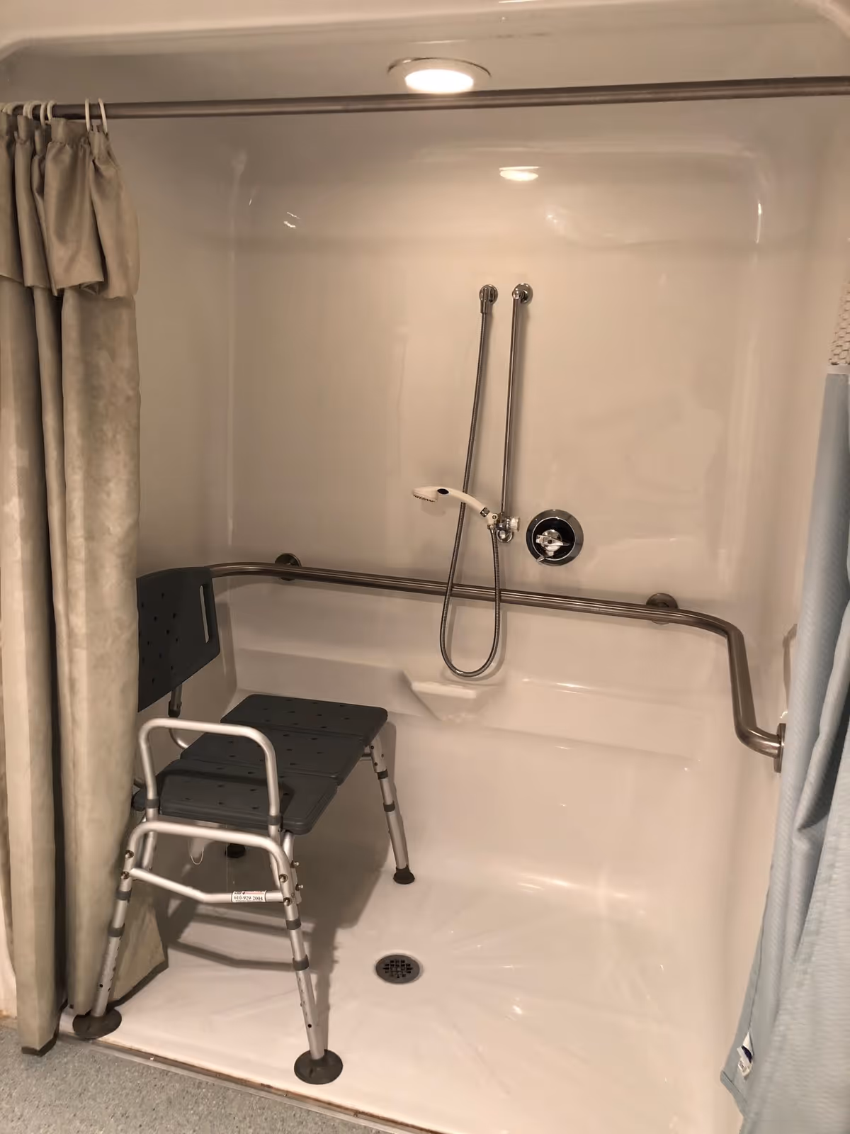 A shower area with a gray shower chair, a handheld showerhead attached to the wall, and a horizontal grab bar for support. The shower is enclosed by beige and light blue curtains.