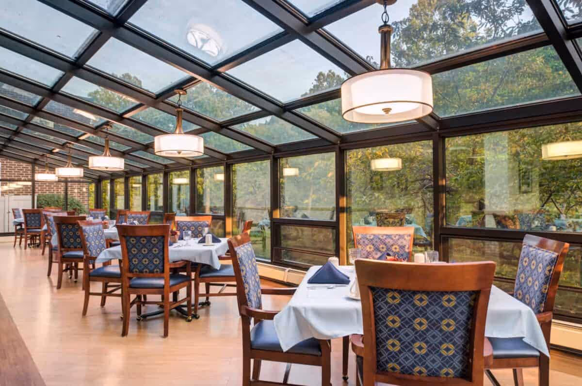 Dining area with multiple tables covered in white tablecloths and set with napkins and utensils. The room has large glass windows and a glass ceiling, allowing natural light to fill the space and providing a view of trees outside. Wooden chairs with patterned blue upholstery surround the tables, and modern pendant lights hang from the ceiling.
