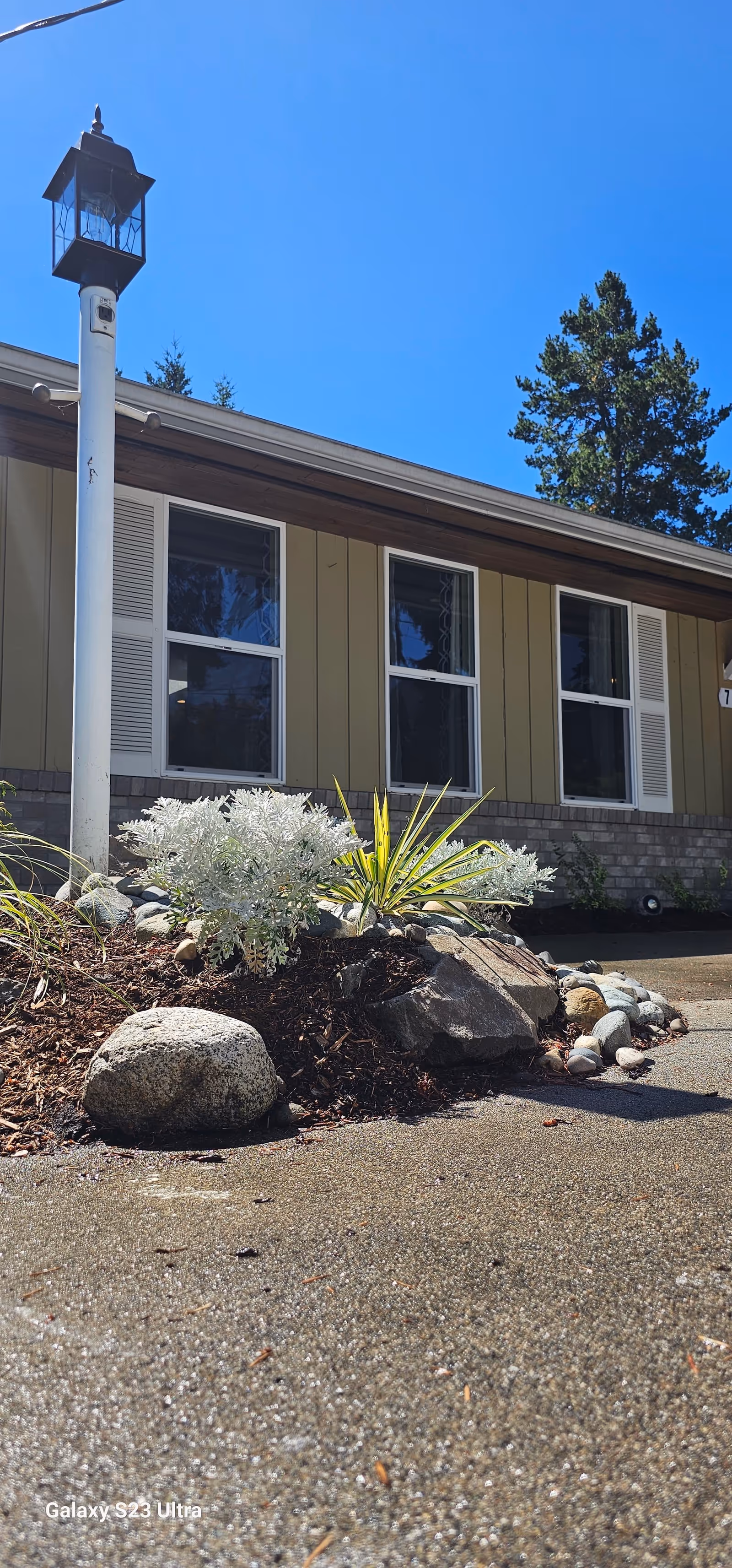Front exterior of a single-story building with windows, a lamp post, and a small rock garden under a clear blue sky.