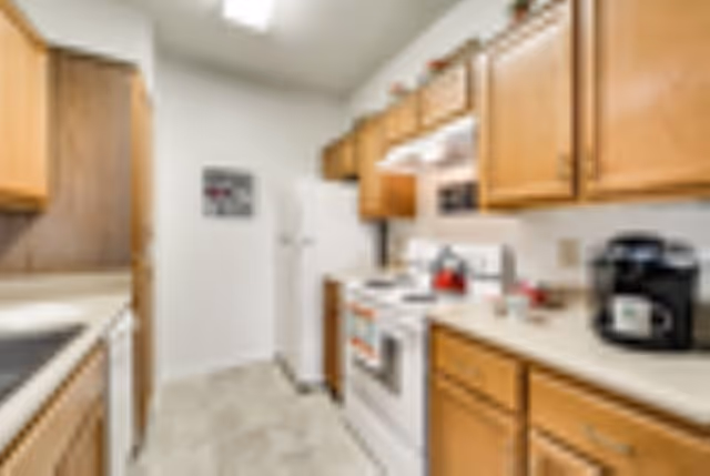 A compact kitchen with wooden cabinets, a white refrigerator, a white stove with a red kettle on it, a coffee maker on the counter, and a dishwasher. The floor is tiled, and the walls are white with a small framed picture hanging.