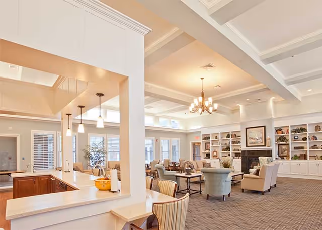 Spacious, well-lit senior community common room with seating areas, built-in bookshelves and fireplace, and a kitchenette/bar counter in the foreground.