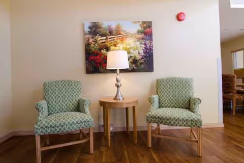 Two patterned green armchairs flank a small round wooden table with a lamp beneath a colorful landscape painting in a seating area.