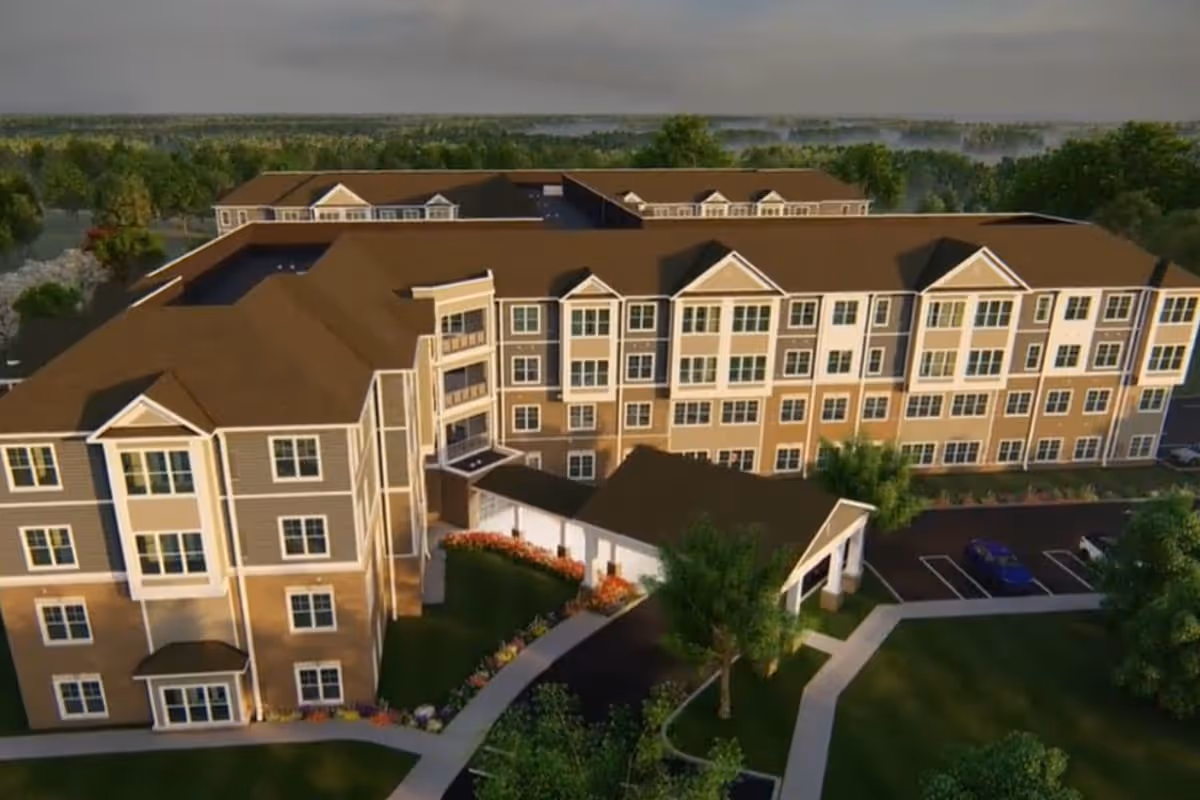 Aerial view of a large, multi-story senior living facility building with brown roofs and beige and gray exterior walls, surrounded by green trees and landscaped gardens, with a parking area visible on the right side.