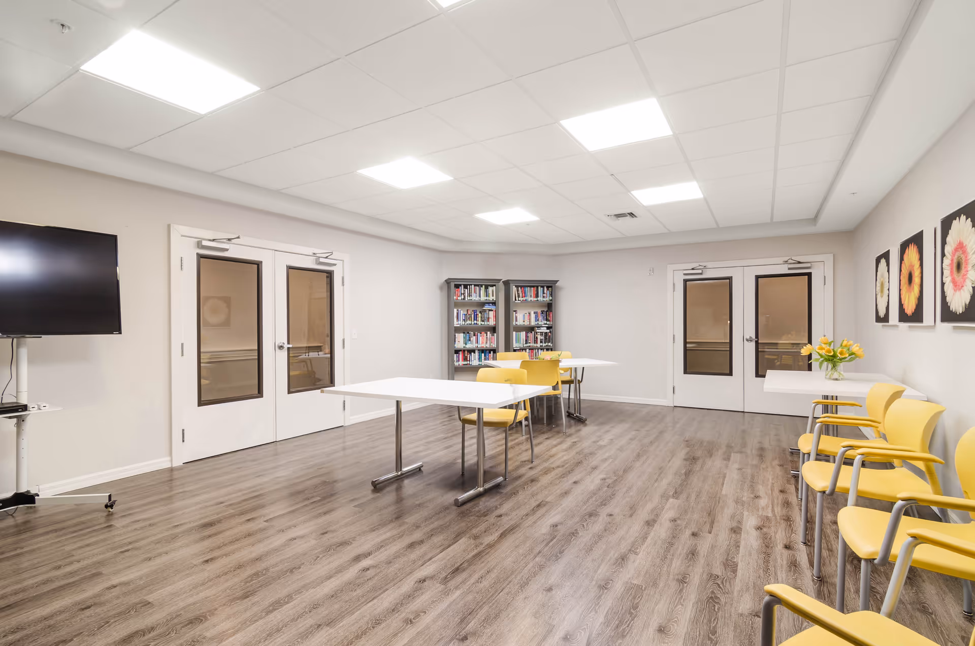 A bright, spacious room with wood flooring and white walls, featuring two white tables with yellow chairs around them. There are bookshelves filled with books against the far wall, a flat-screen TV mounted on a stand to the left, and three framed flower pictures on the right wall. Double doors with windows are visible on both the left and right sides of the room.
