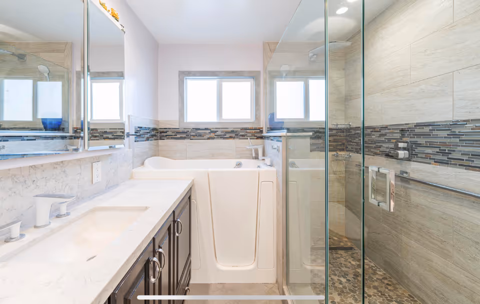Bright bathroom featuring a walk-in bathtub with a door, a glass-enclosed shower with pebble stone flooring, a marble countertop with a sink and faucet, and a large mirror above the sink. The walls are tiled with beige tiles and a decorative horizontal strip of mosaic tiles. A window provides natural light.