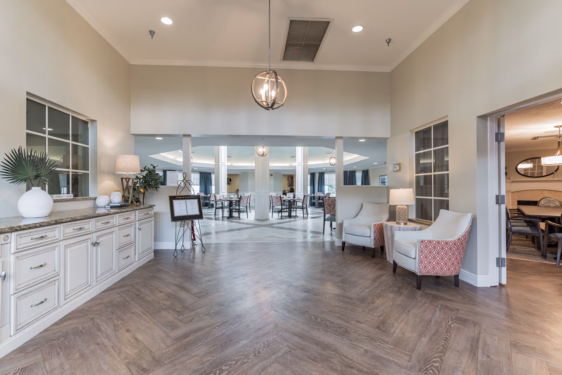 A spacious and well-lit interior area of a senior living facility with wooden flooring, white cabinetry on the left, two armchairs with a small table and lamp on the right, and a dining area visible through a large open doorway in the background. The walls are painted light beige, and there are modern light fixtures hanging from the ceiling.