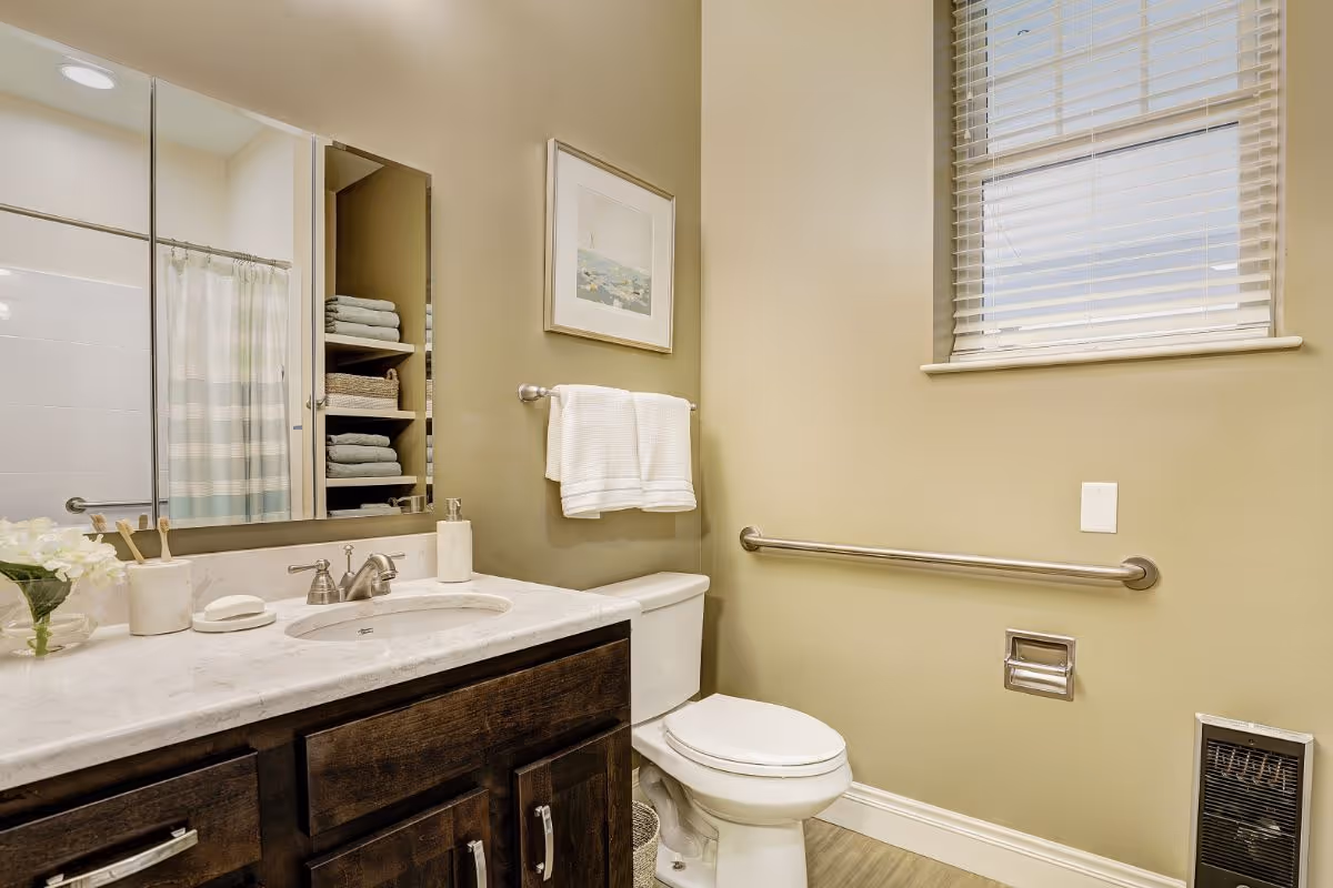 A clean and well-lit bathroom featuring a white toilet with a safety grab bar on the wall beside it, a dark wood vanity with a marble countertop and an under-mount sink, a large mirror above the sink, a towel rack with two white towels, a window with blinds, and shelves stocked with folded towels and baskets.