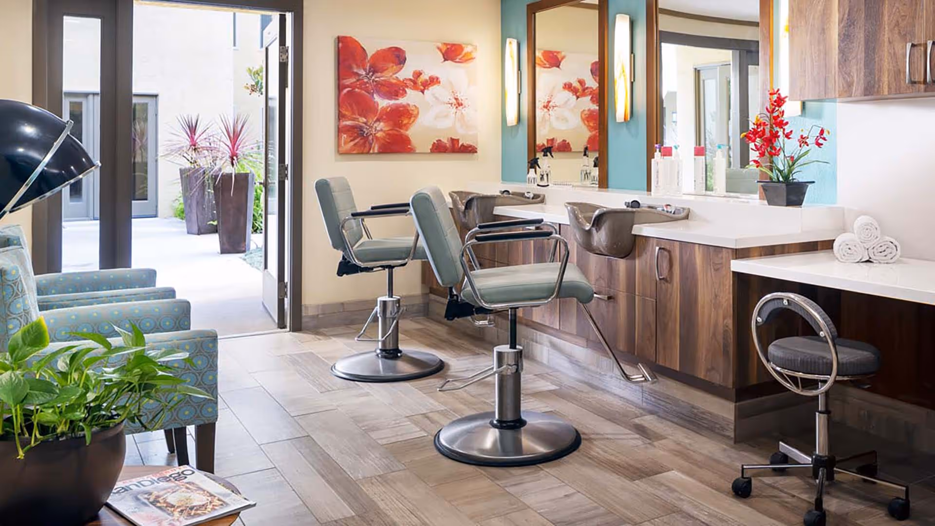 Interior view of a salon area in a senior living facility with two salon chairs in front of a counter with sinks and mirrors. There are decorative red flower paintings on the wall, a small potted plant on the counter, and a seating area with patterned armchairs and a floor lamp near an open door leading outside.