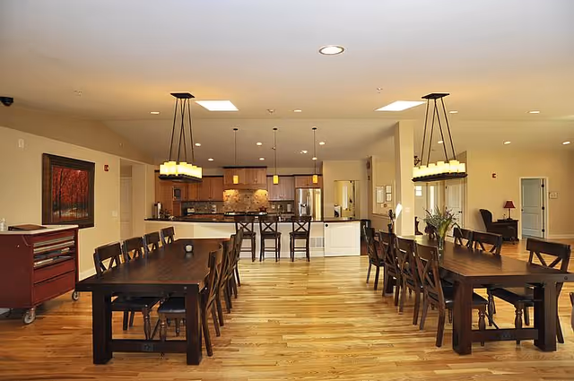 A spacious dining area with two long wooden tables surrounded by chairs. The room features wooden flooring, modern pendant lights hanging from the ceiling, and a kitchen area in the background with stainless steel appliances and wooden cabinets.