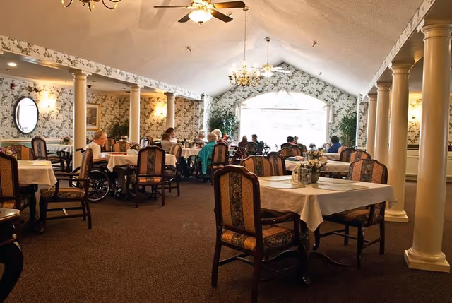 Interior view of a dining room in a senior living facility with several tables covered with white tablecloths and chairs around them. Elderly residents are seated at some tables, some in wheelchairs. The room has floral wallpaper, carpeted floor, ceiling fans, chandeliers, and large windows letting in natural light.