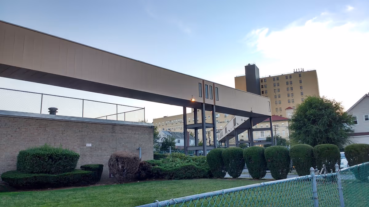 Exterior view of a nursing facility with an elevated covered walkway connecting buildings, trimmed shrubs, and a chain-link fence.