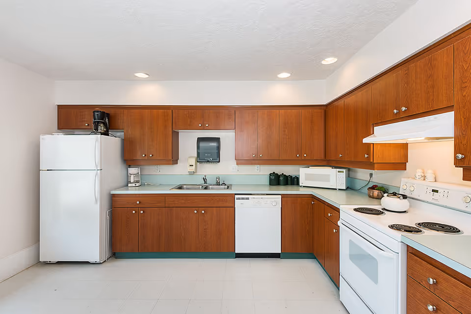 A clean and well-lit kitchen with wooden cabinets, a white refrigerator, a white stove with four burners, a white microwave, a dishwasher, a coffee maker, and a double sink. The countertops are light-colored, and there is a paper towel dispenser mounted on the wall above the sink.