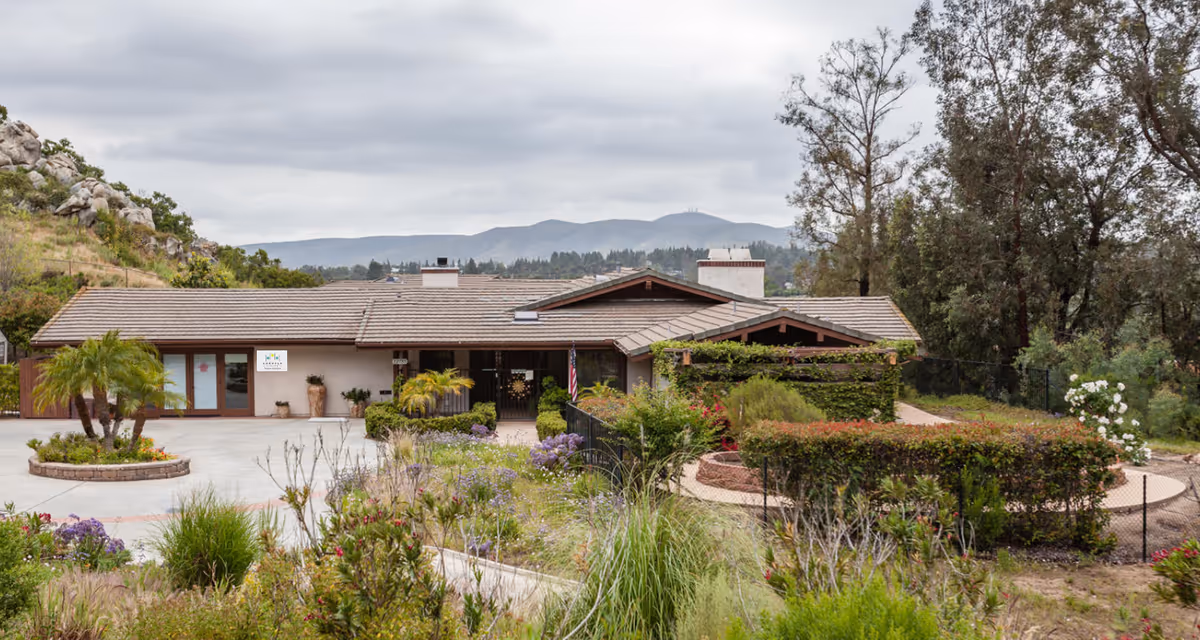 Single-story senior living building with a tiled roof, landscaped gardens and circular driveway against a backdrop of hills.