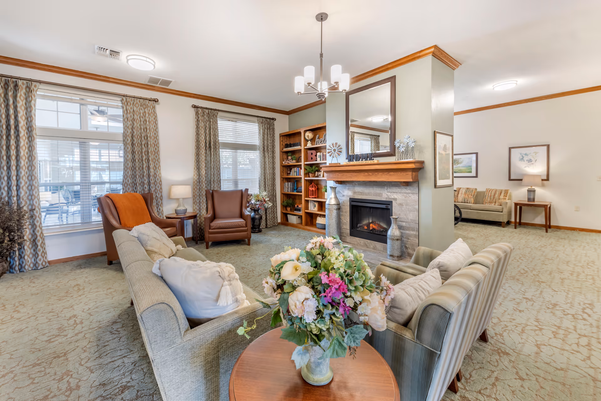 Bright furnished common room with sofas and armchairs arranged around a fireplace and a floral centerpiece on a table.