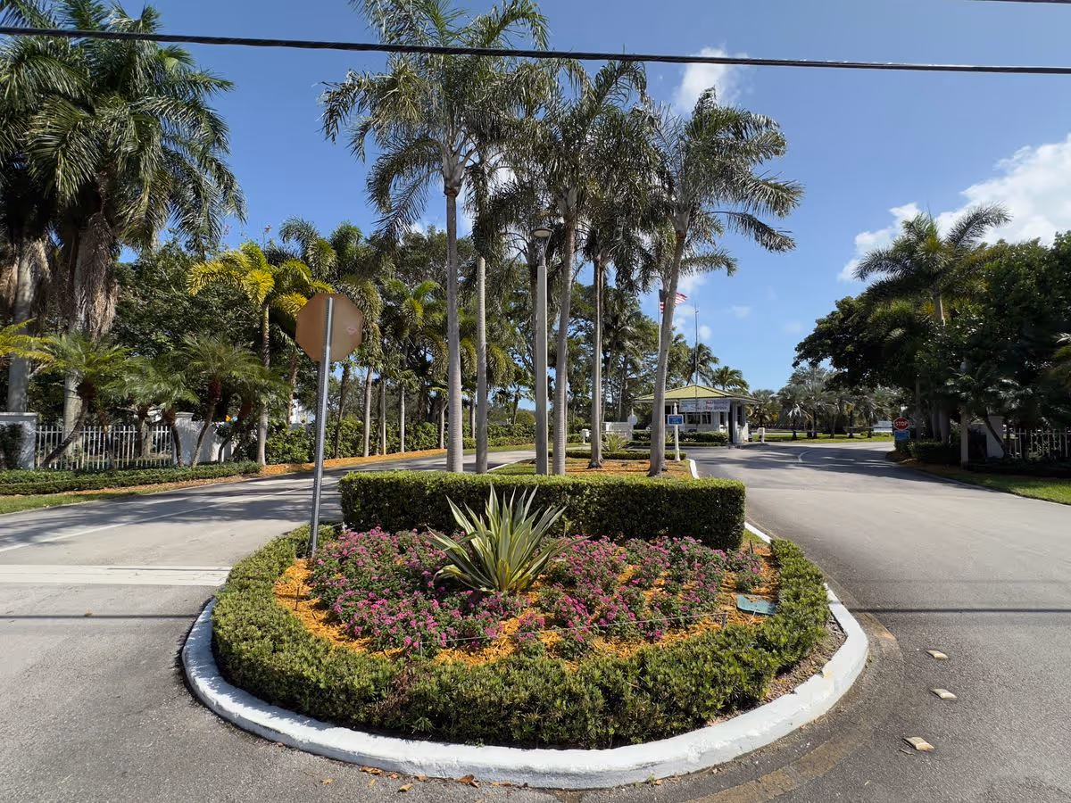 Landscaped driveway entrance with a circular flowerbed, palm trees, and a guardhouse under a blue sky.