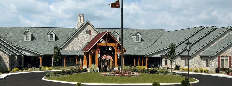 Front exterior view of a large stone building with a gray shingled roof and wooden entrance structure, surrounded by landscaped greenery and a circular driveway with a flagpole in the center.