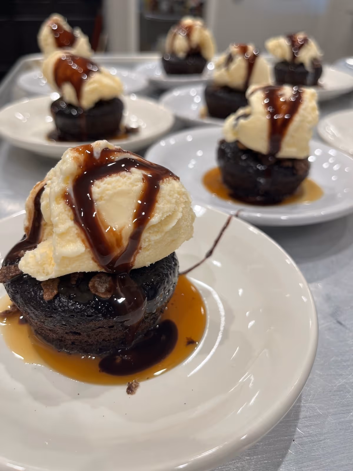Close-up view of several small chocolate cakes topped with vanilla ice cream and drizzled with chocolate syrup, each served on a white plate.