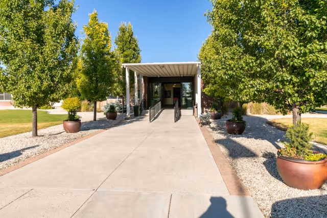 Covered entrance with wheelchair ramps leading to a building, flanked by trees and large potted planters.