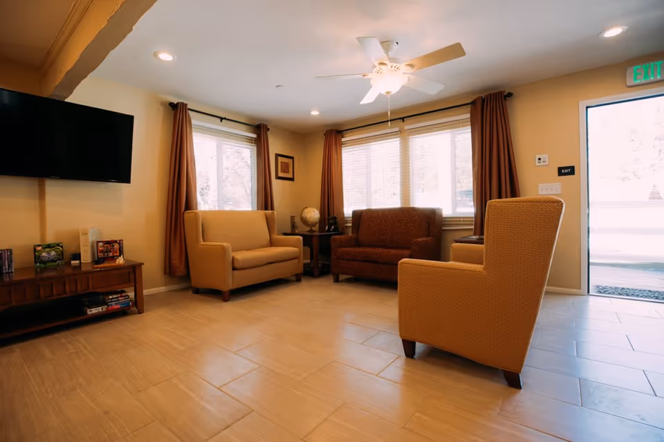 A cozy living room area with three armchairs and a loveseat arranged around a corner. The room has beige walls, large windows with brown curtains, a ceiling fan with light, a wall-mounted flat-screen TV, and a wooden media console with books and games. An exit door with a green exit sign is visible on the right side.