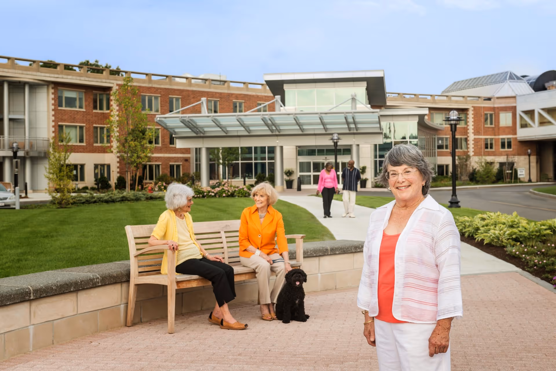 Three elderly women outside a modern retirement community building. Two women are sitting on a wooden bench with a small black dog, while another woman stands smiling in the foreground. In the background, a couple walks on the pathway leading to the building entrance surrounded by green lawns and landscaping.