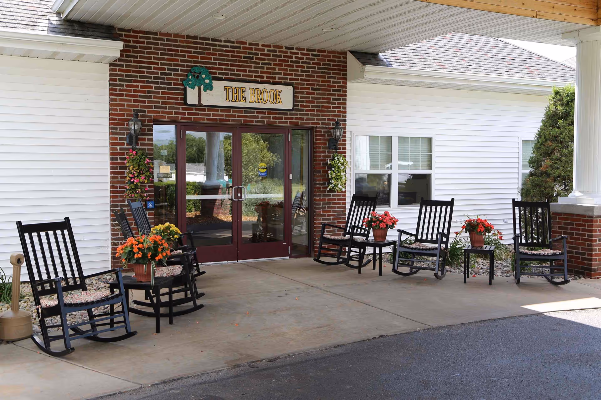 Covered entrance to The Brook with double glass doors, a sign reading "THE BROOK", rocking chairs and potted flowers on the porch.