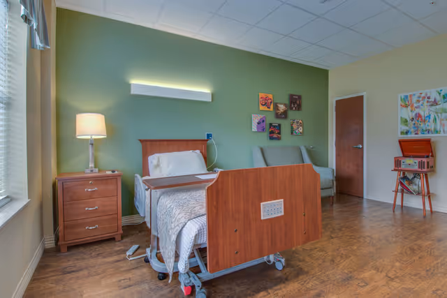 A bright patient bedroom with a medical bed, wooden nightstand and lamp, an armchair and colorful wall art.