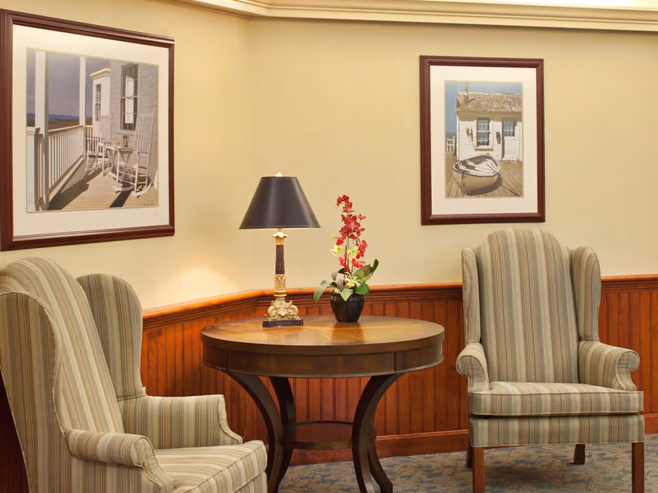 A cozy seating area with two striped upholstered armchairs facing a round wooden table. On the table, there is a decorative lamp with a black shade and a vase with red and white flowers. The walls are light-colored with wooden wainscoting, and two framed pictures depicting outdoor scenes hang above the chairs.
