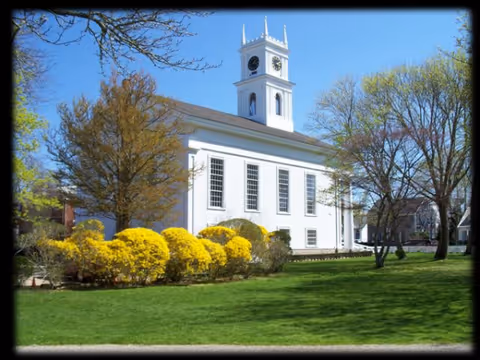 White building with clock tower sits on a green lawn with trees.