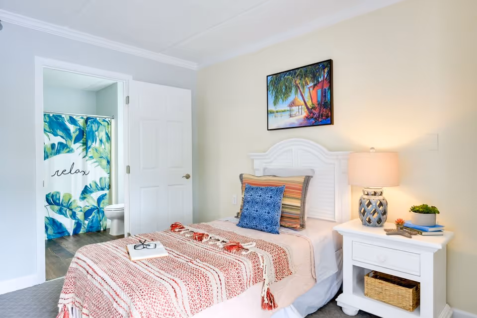 Bright single bedroom with a made bed, nightstand and lamp, colorful artwork above the headboard, and an open bathroom featuring a tropical 'relax' shower curtain.