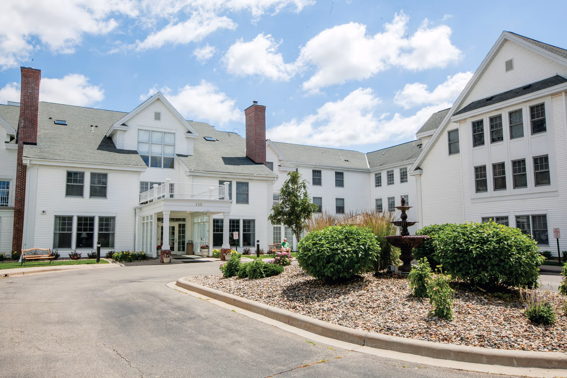 Exterior view of a senior living facility with white siding, multiple windows, and two brick chimneys. The building surrounds a circular driveway with landscaped bushes, a small tree, and a tiered water fountain in the center. The sky is partly cloudy with blue patches visible.