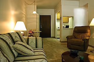 A cozy living room area with a patterned sofa and matching cushions on the left, a brown recliner chair on the right, a wooden coffee table in front, and a lamp on a side table providing warm lighting. In the background, there is a small dining table with chairs, a dark wooden door, and a partial view of a kitchen area with light-colored cabinets.