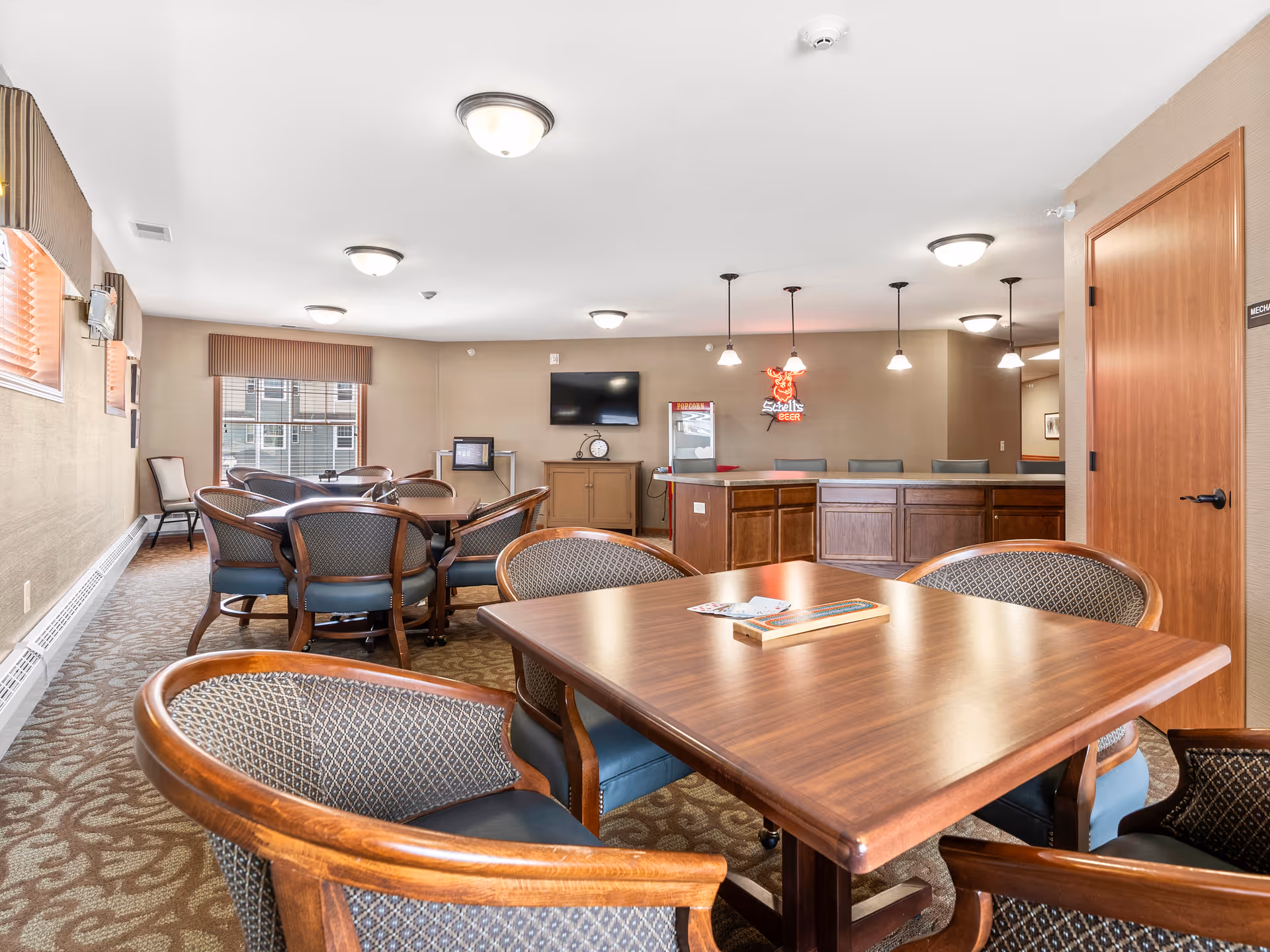 Bright communal dining and lounge area with wooden tables and upholstered chairs, a TV on the far wall and a counter/bar area with pendant lights.