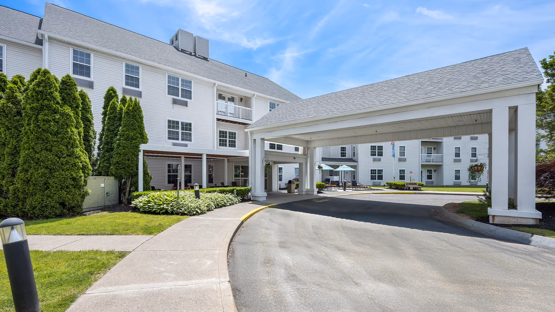 Exterior view of Holiday Lodge at Cold Spring showing a covered driveway entrance, white multi-story building with multiple windows, green landscaping including tall bushes and grass, and a clear blue sky.