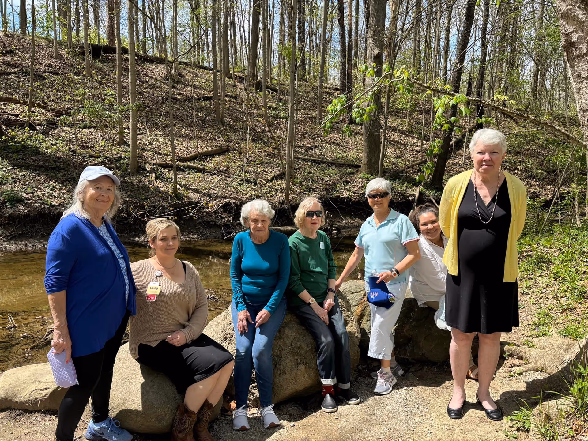 A group of seven women, mostly elderly, posing outdoors in a wooded area near a small creek. Some are sitting on large rocks while others stand nearby. The women are dressed casually and appear to be enjoying a sunny day in nature.