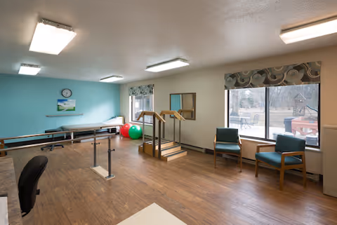 A rehabilitation room with wooden flooring and light blue walls. The room contains parallel bars for walking exercises, a small set of stairs with handrails, two exercise balls, two chairs near large windows, and a clock on the wall above a bench.