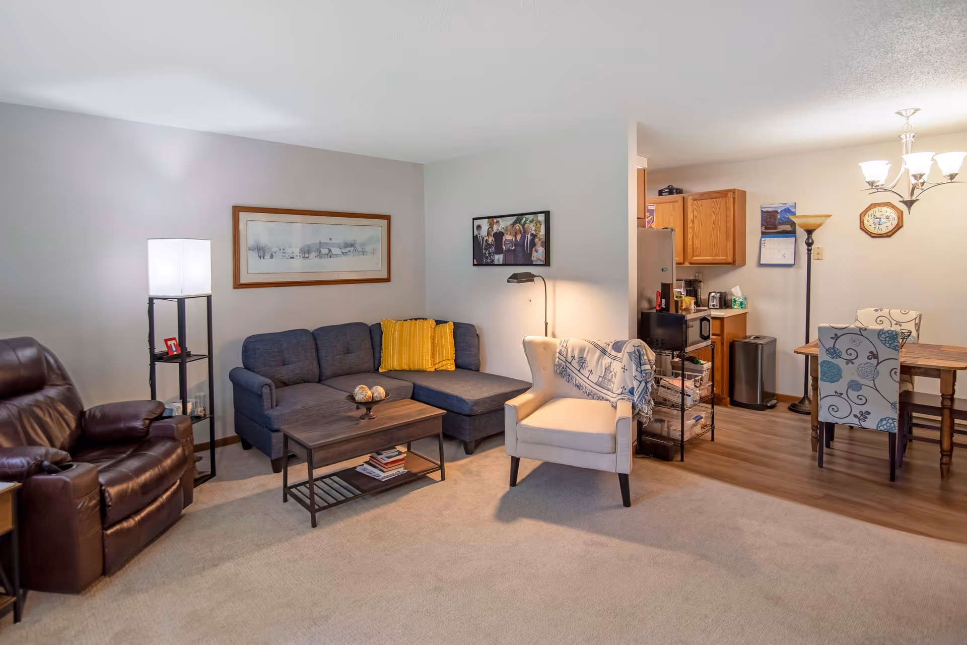 A cozy senior living room with a dark blue sectional sofa adorned with a yellow pillow, a beige armchair with a blue and white throw blanket, and a brown leather recliner. A wooden coffee table with decorative items sits in the center on a beige carpet. The room has soft lighting from a floor lamp and a standing lamp with shelves. In the background, there is a small dining area with a wooden table and floral-patterned chairs, a kitchen with wooden cabinets, and a wall clock. The walls are decorated with framed pictures and a calendar.