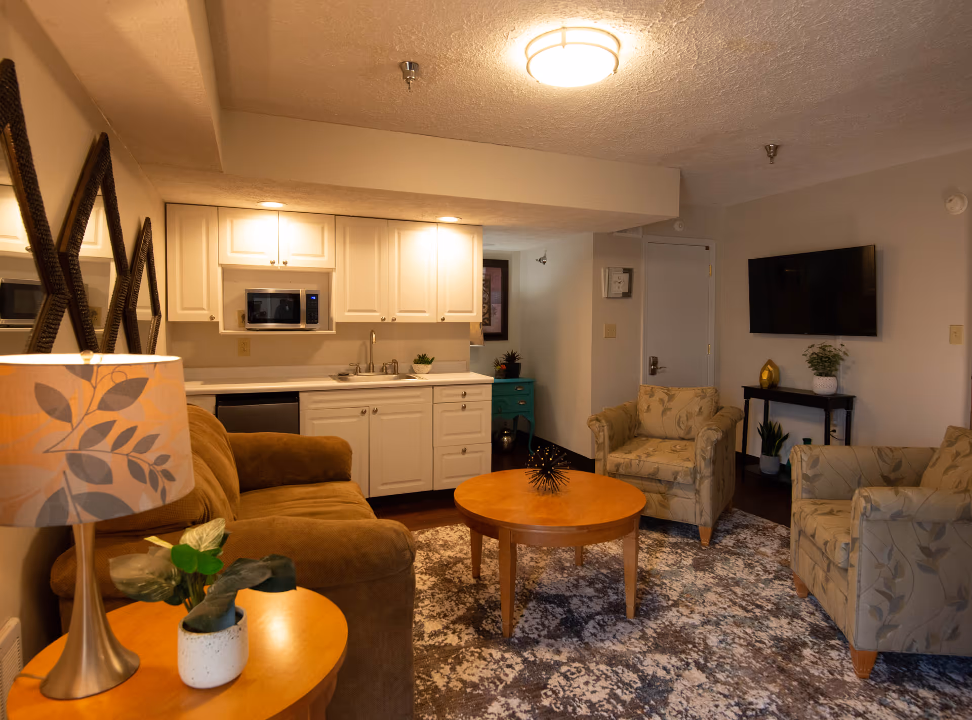 A cozy living room area with a brown sofa, two patterned armchairs, a round wooden coffee table, and a side table with a lamp and a small plant. The room has a patterned rug, a wall-mounted TV, and a small kitchenette with white cabinets, a microwave, and a sink in the background.