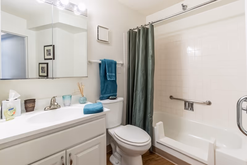 A clean bathroom featuring a white vanity with a sink, a large mirror with lights above, a toilet with a teal towel hanging on a bar above it, and a bathtub with a green shower curtain and a safety grab bar installed on the tiled wall.