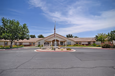 Front exterior of a single-story senior living building with a flagpole and circular driveway under a partly cloudy sky.